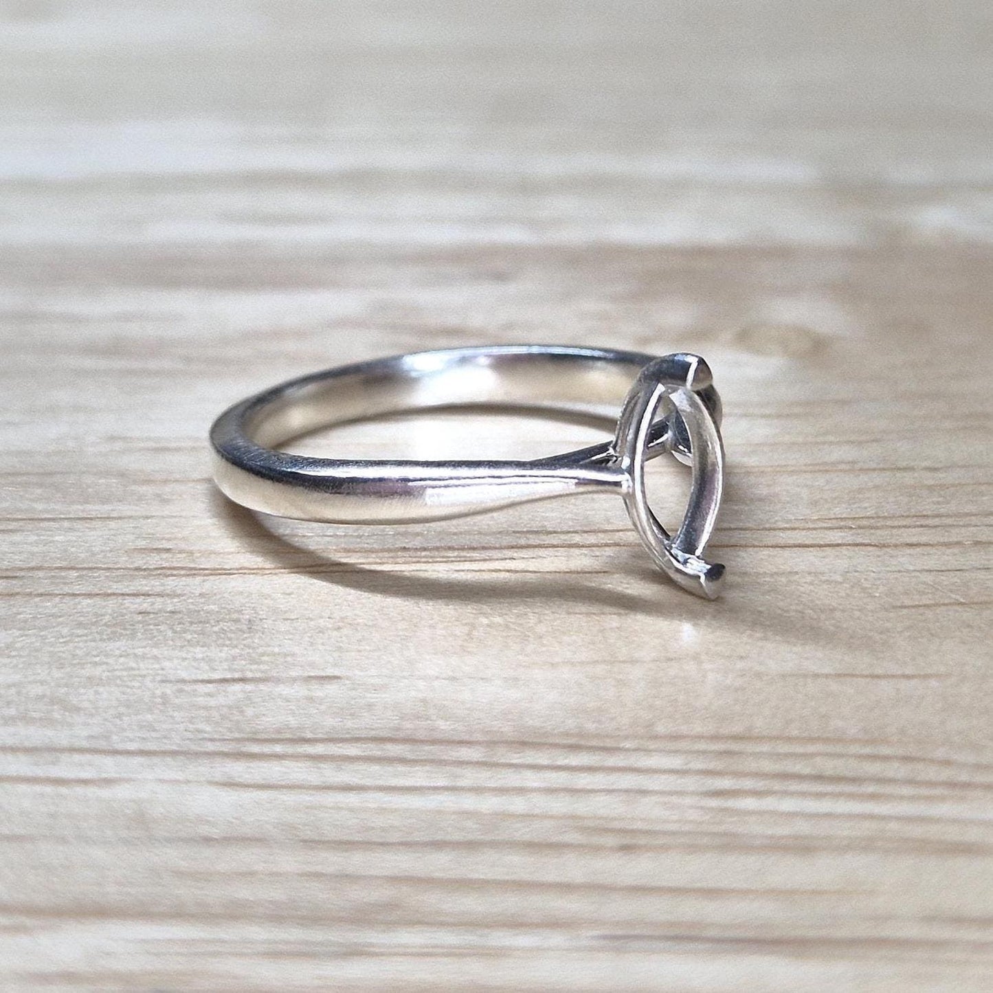 Side view of a delicate silver ring blank with cathedral-style shoulders and marquise setting, set on a pale wood background.