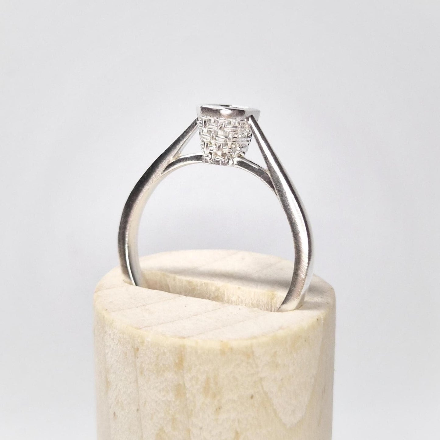 Side profile of a silver ring blank with an open basket bezel setting, displayed upright on a wooden base with a white background.