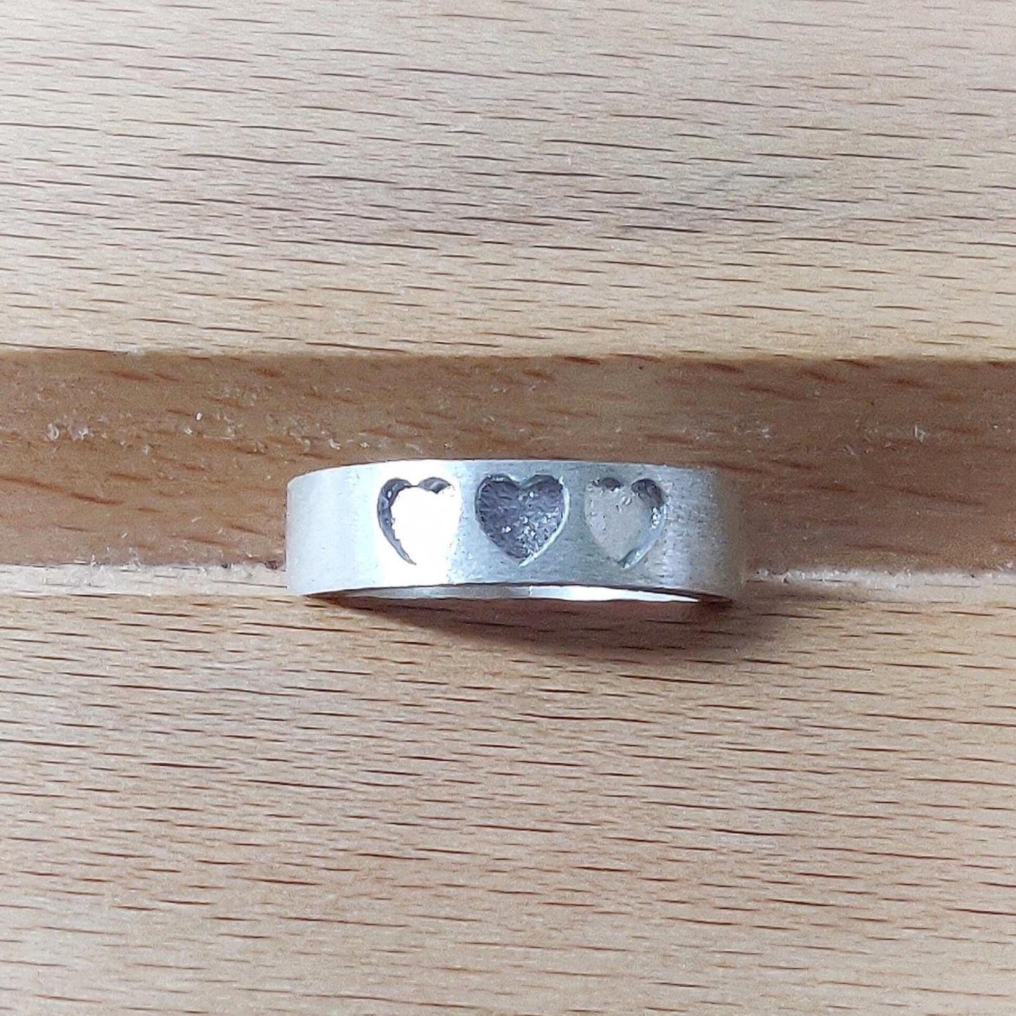 Brushed silver keepsake ring with three heart inlays, photographed on a pale wooden surface.