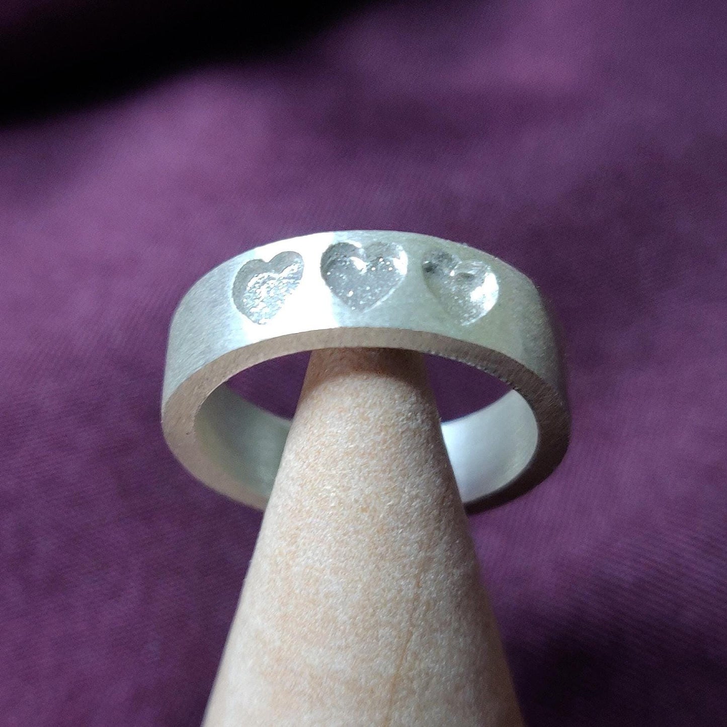 Silver keepsake ring worn on a hand, showcasing three recessed heart shapes against a soft purple cloth.