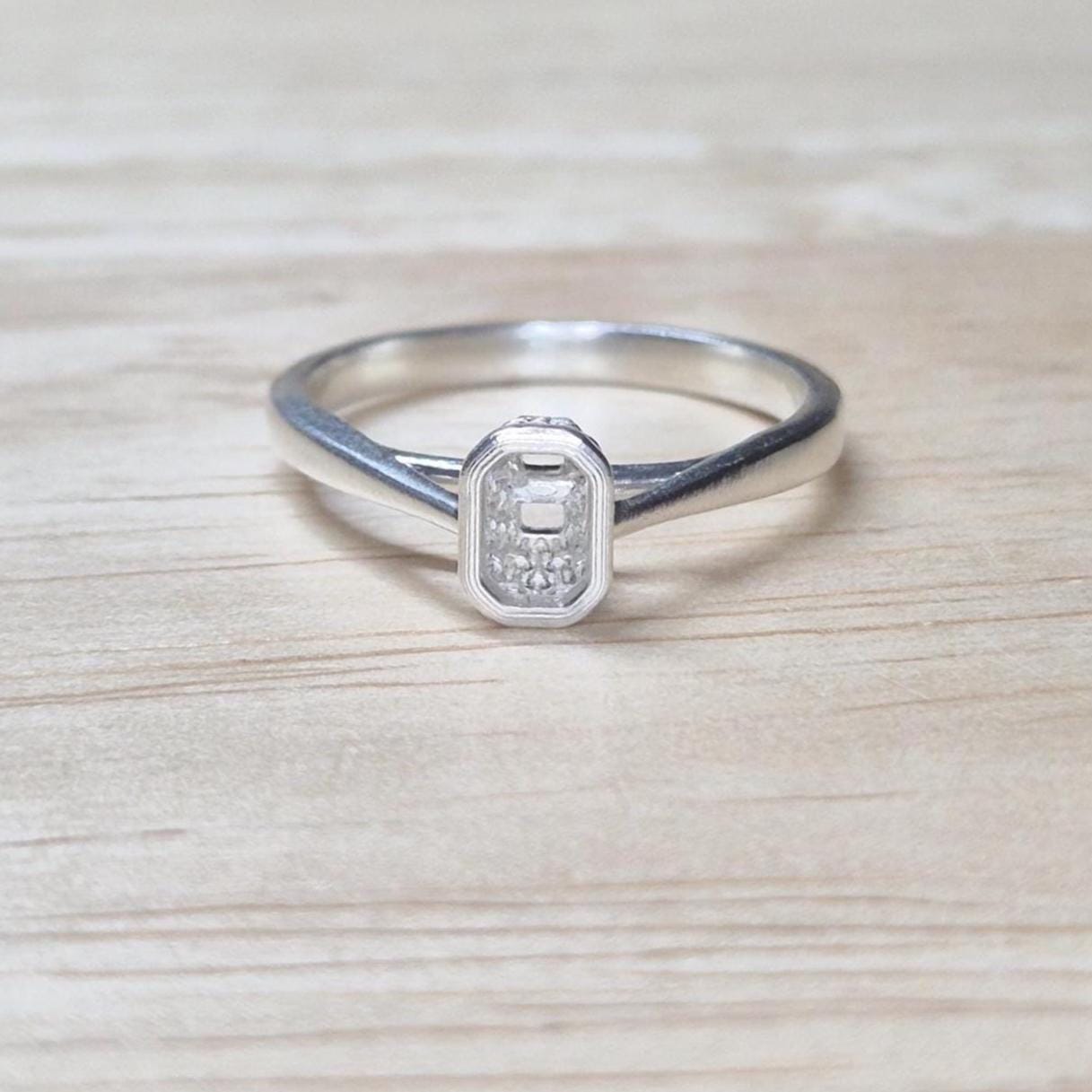 Straight-on view of silver ring blank with rectangular bezel setting, photographed against a wood background with neutral tones.