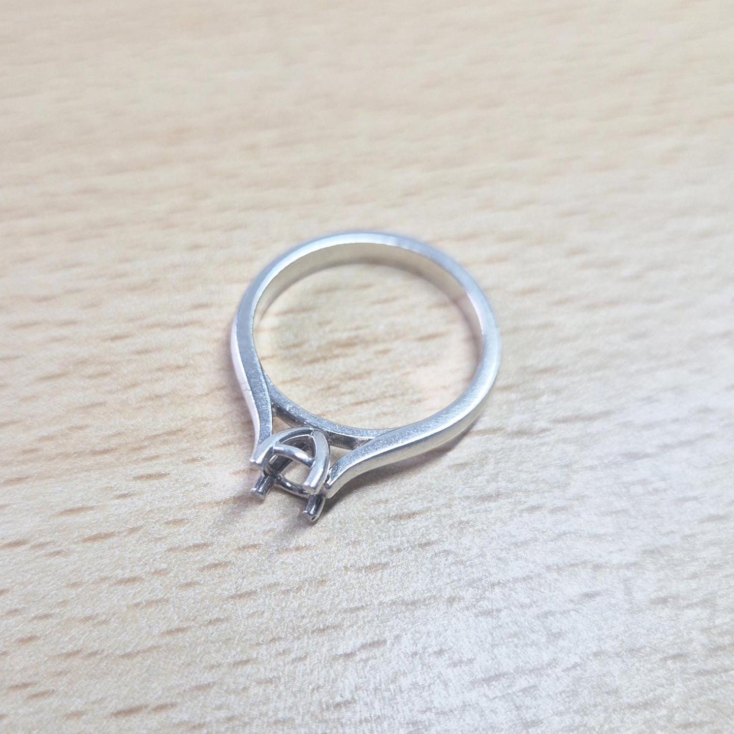 Sterling silver ring setting with a four-prong round head, resting on a light wood surface, showing the open basket-style setting and the complete ring design from above.