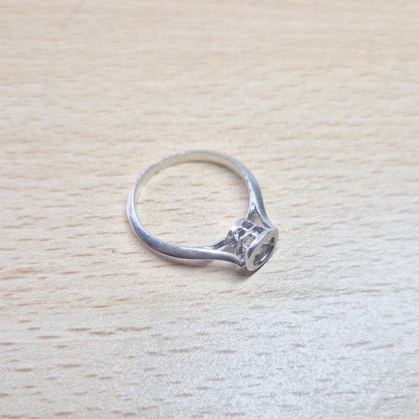 A silver ring setting with an empty, ornate bezel featuring small cut-out details, lying on a light wooden surface. The ring is viewed from a slight angle, showcasing its side profile.