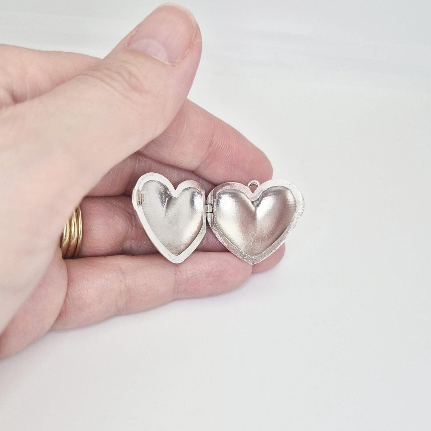 A hand holding an open, puffed silver heart locket. The two heart halves are spread apart, revealing their empty interiors, with a small loop for a chain on one side. Gold rings are visible on the fingers.