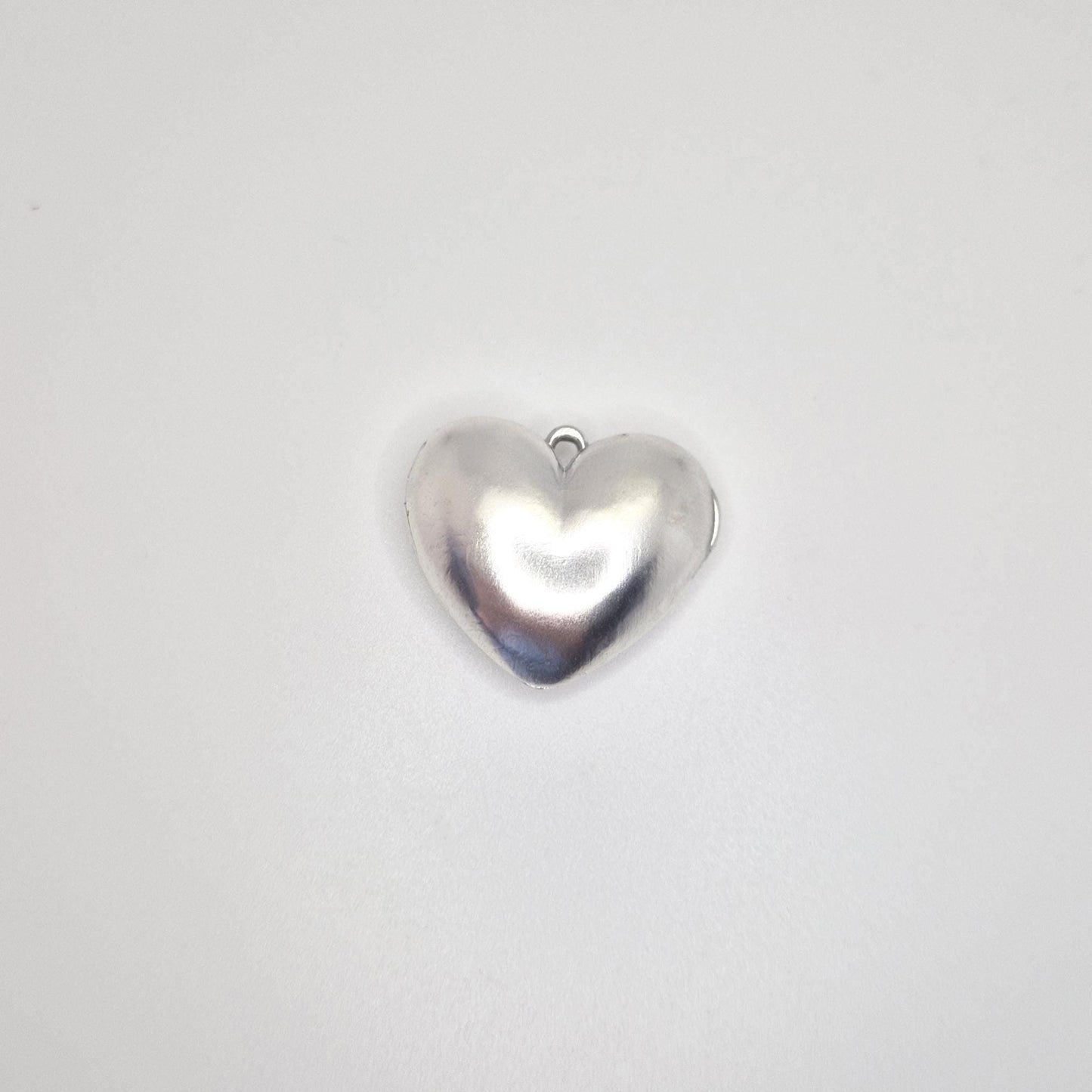 A shiny, closed, puffed silver heart locket with a small loop at the top, presented against a clean white background. The front of the locket is visible, showing its smooth, curved shape.