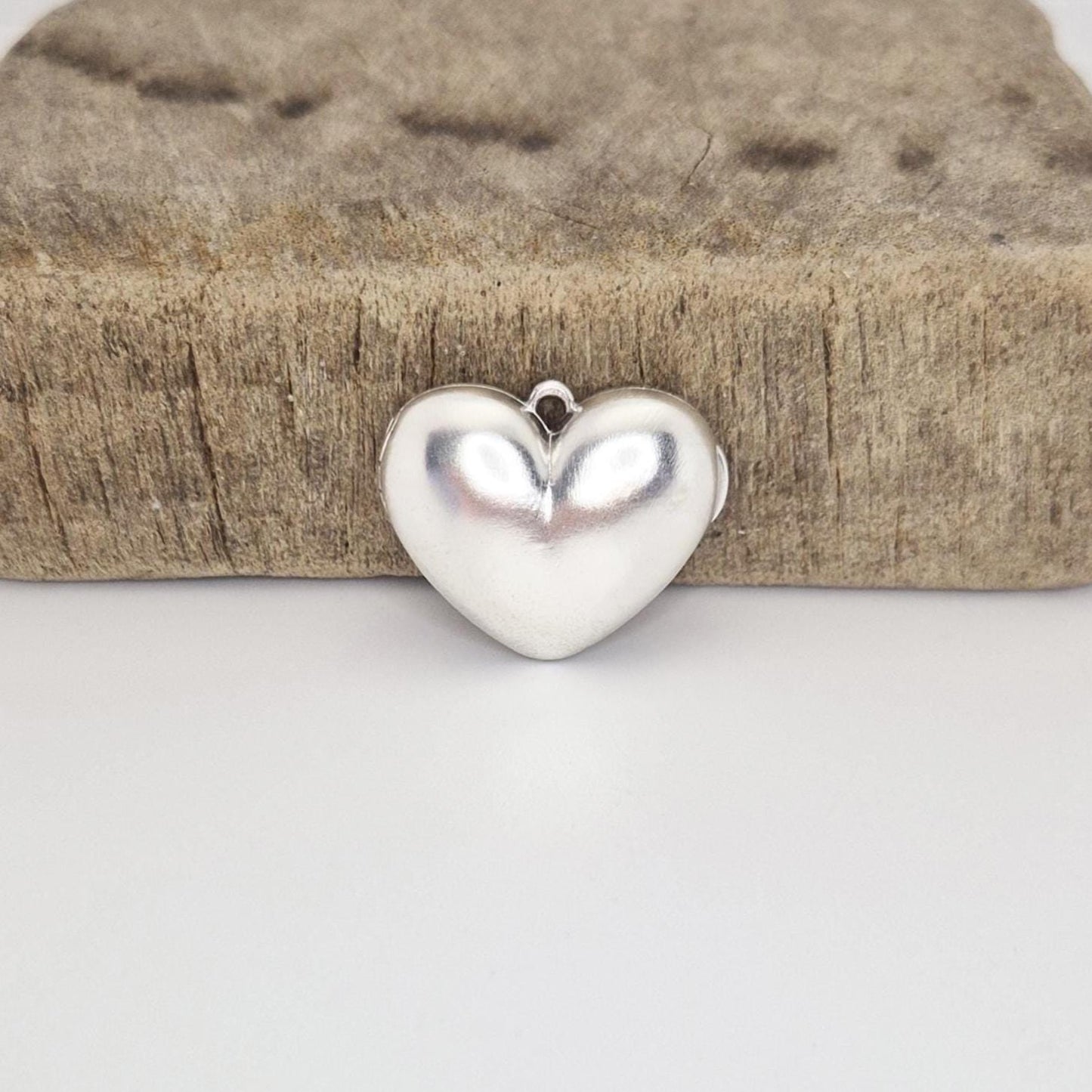 A closed, puffed silver heart locket positioned in front of a rustic wooden block on a white surface. The locket's reflective surface and the small attachment loop are clearly visible.