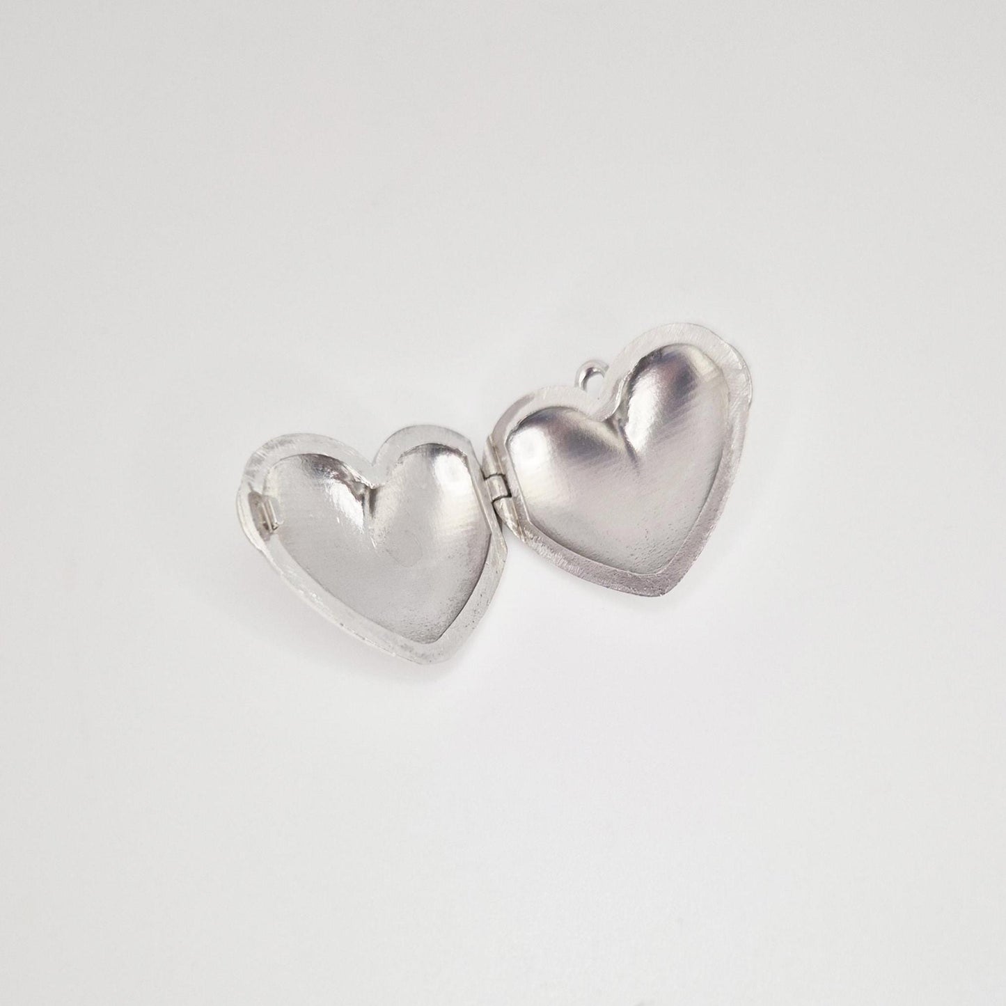 An open, puffed silver heart locket against a plain white background. The two halves of the heart are spread open, showing the smooth, concave interior of each side, with a small jump ring visible on the right half.