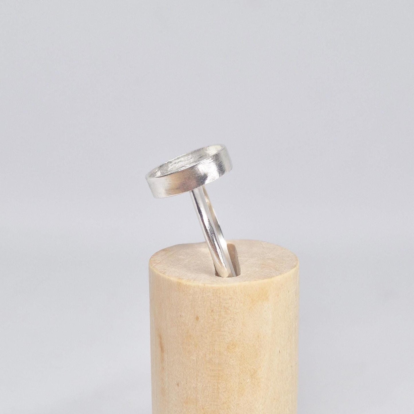 Side view of an empty sterling silver oval cabochon ring setting, standing upright on a light wood cylindrical display.