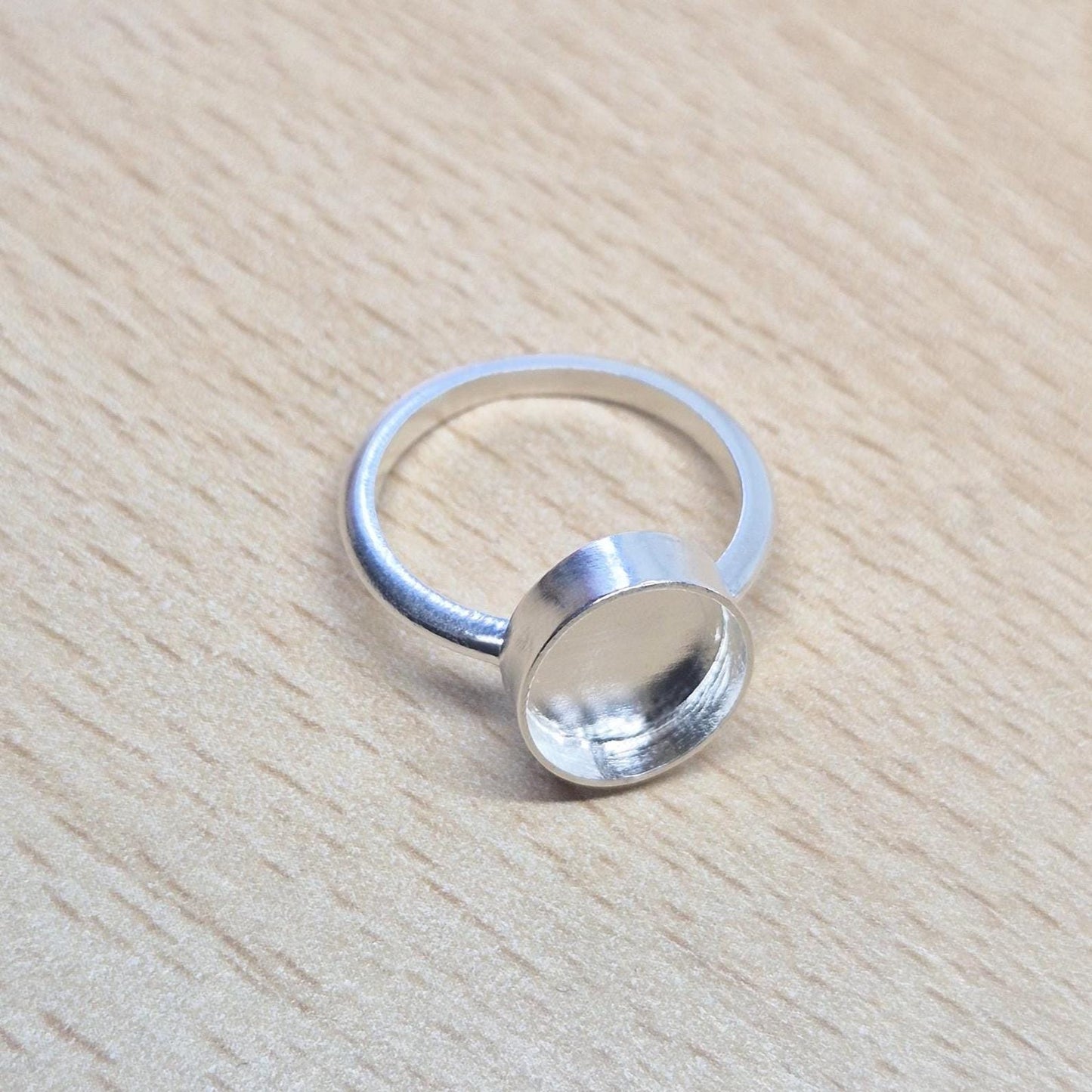 Top-down view of a sterling silver oval cabochon ring setting resting on a light wood surface, showcasing the empty bezel.