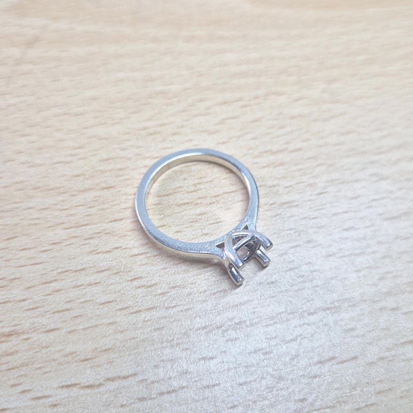 Sterling silver ring setting with a four-prong square head, resting on a light wood surface, providing a clear, slightly elevated view of the entire ring.