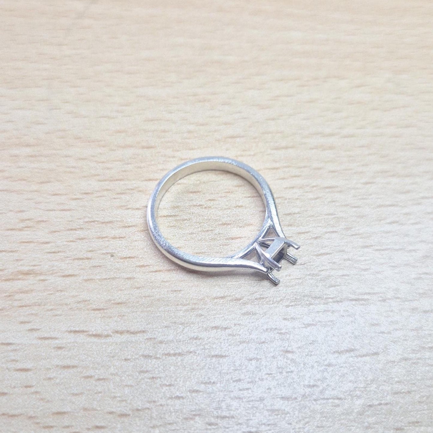 Sterling silver ring setting with a four-prong princess cut head, resting on a light wood surface, providing an overhead view of the full ring and its open setting.