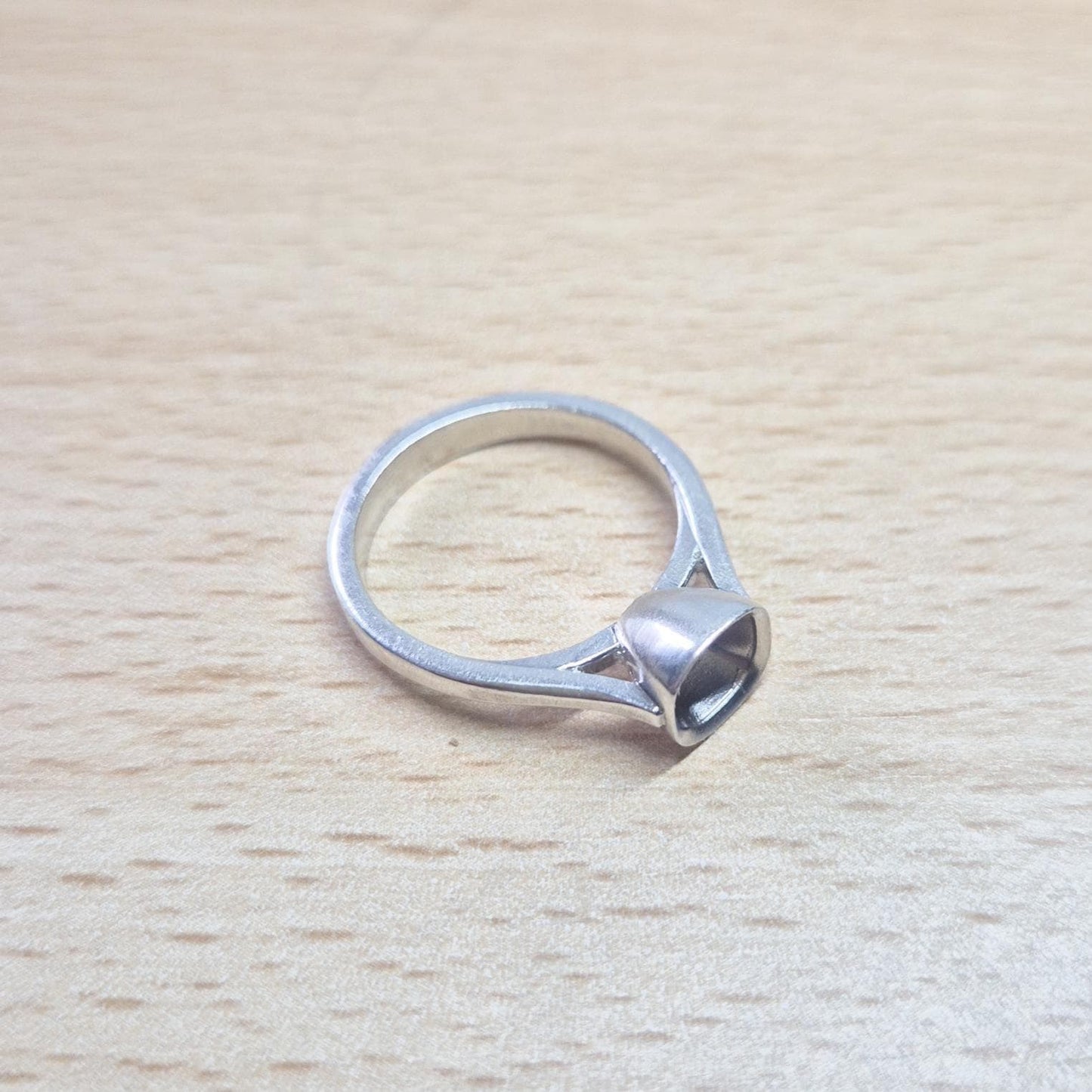 Sterling silver ring setting with a cushion cut bezel, resting on a light wood surface, providing an overhead view of the full ring and the empty bezel setting.