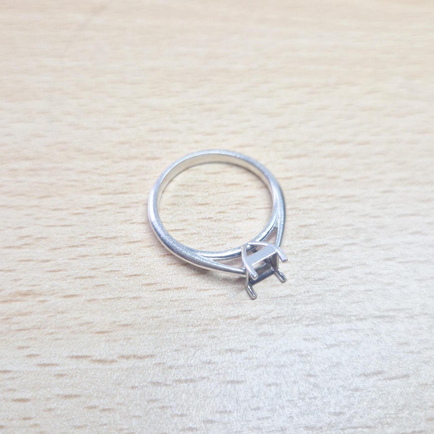 Silver cathedral setting princess cut ring blank lying flat on a light wooden surface, showing the top of the empty four-prong setting and the band.