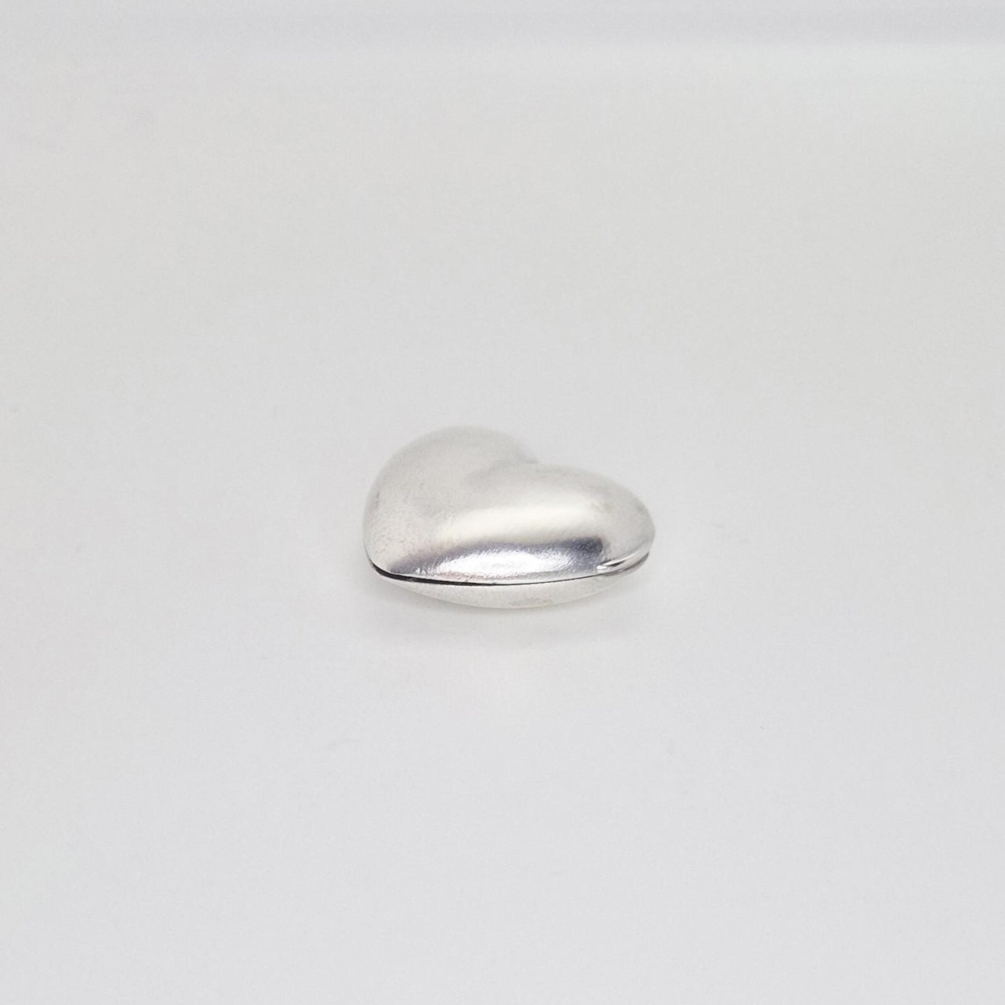 A closed, puffed silver heart locket seen from the back, showing the seam where the two halves join and its rounded contours on a plain white background.