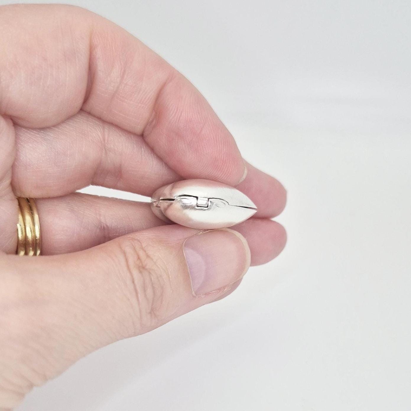 A hand holding a closed, puffed silver heart locket, viewed from the side, emphasizing its plump shape and the smooth hinge where the two halves meet. Gold rings are visible on the fingers.