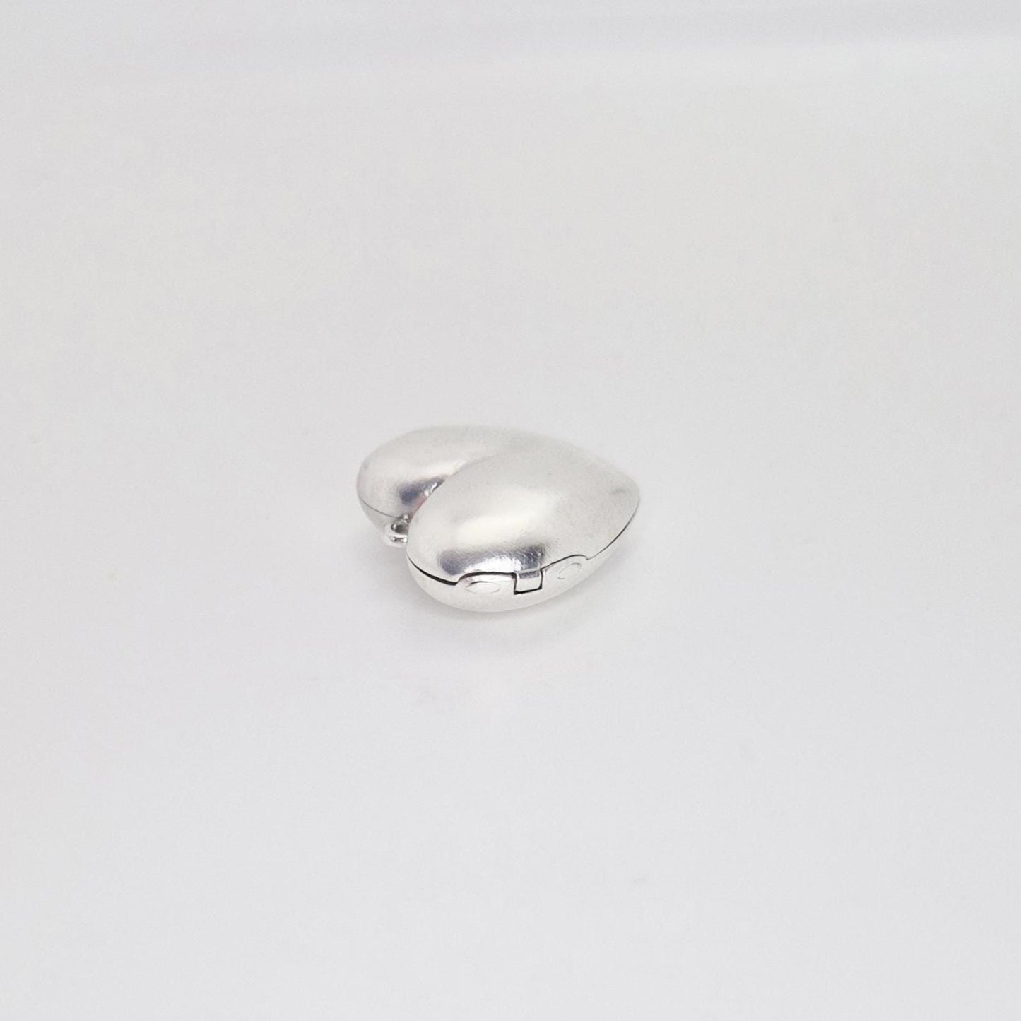 A closed, puffed silver heart locket viewed from the side on a white background, highlighting its thick, rounded profile and the hinge mechanism.