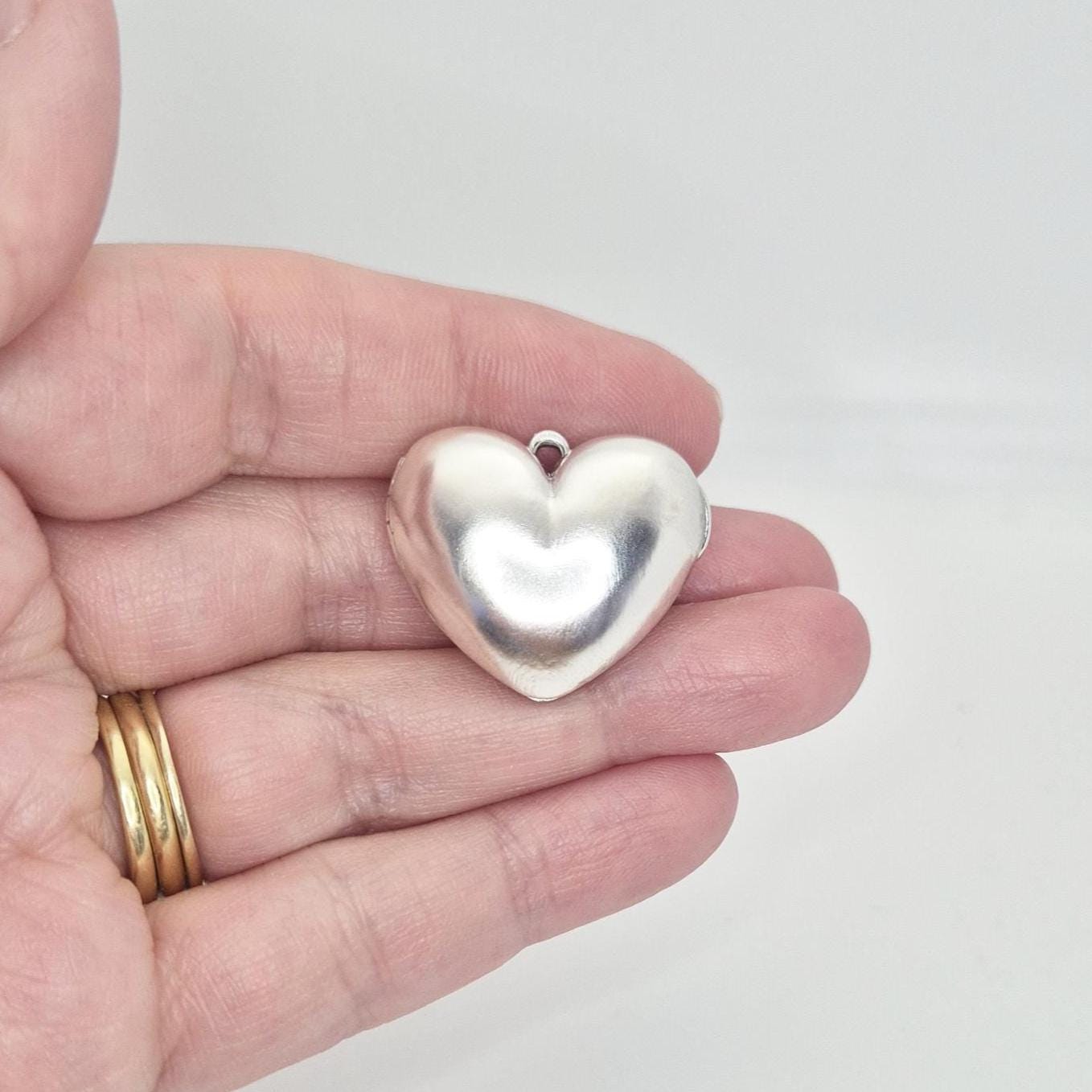 A hand holding a closed, puffed silver heart locket, showcasing its size relative to an adult palm. The locket has a shiny, reflective surface and a small loop for attaching to a chain. Gold rings are visible on the fingers.