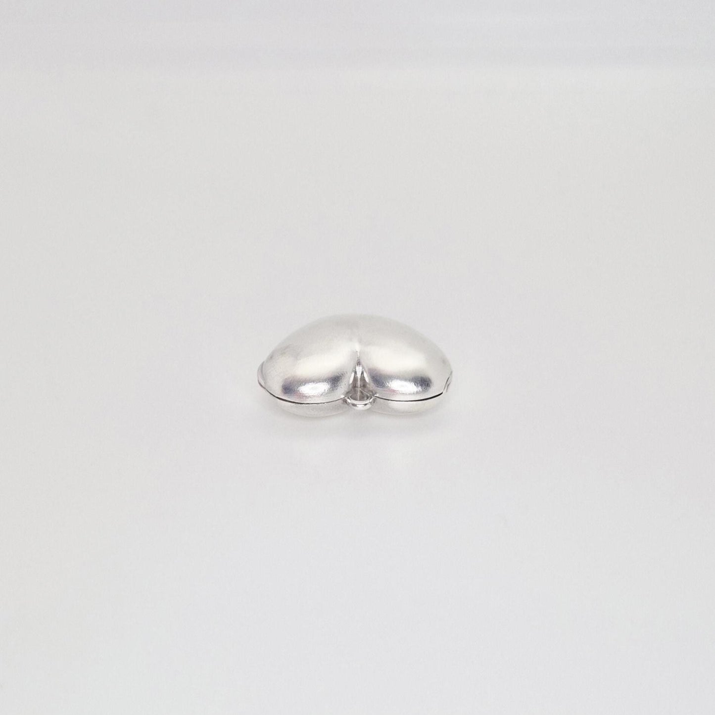 A shiny, puffed silver heart locket, closed and lying on a plain white background, shown from a slightly elevated angle.