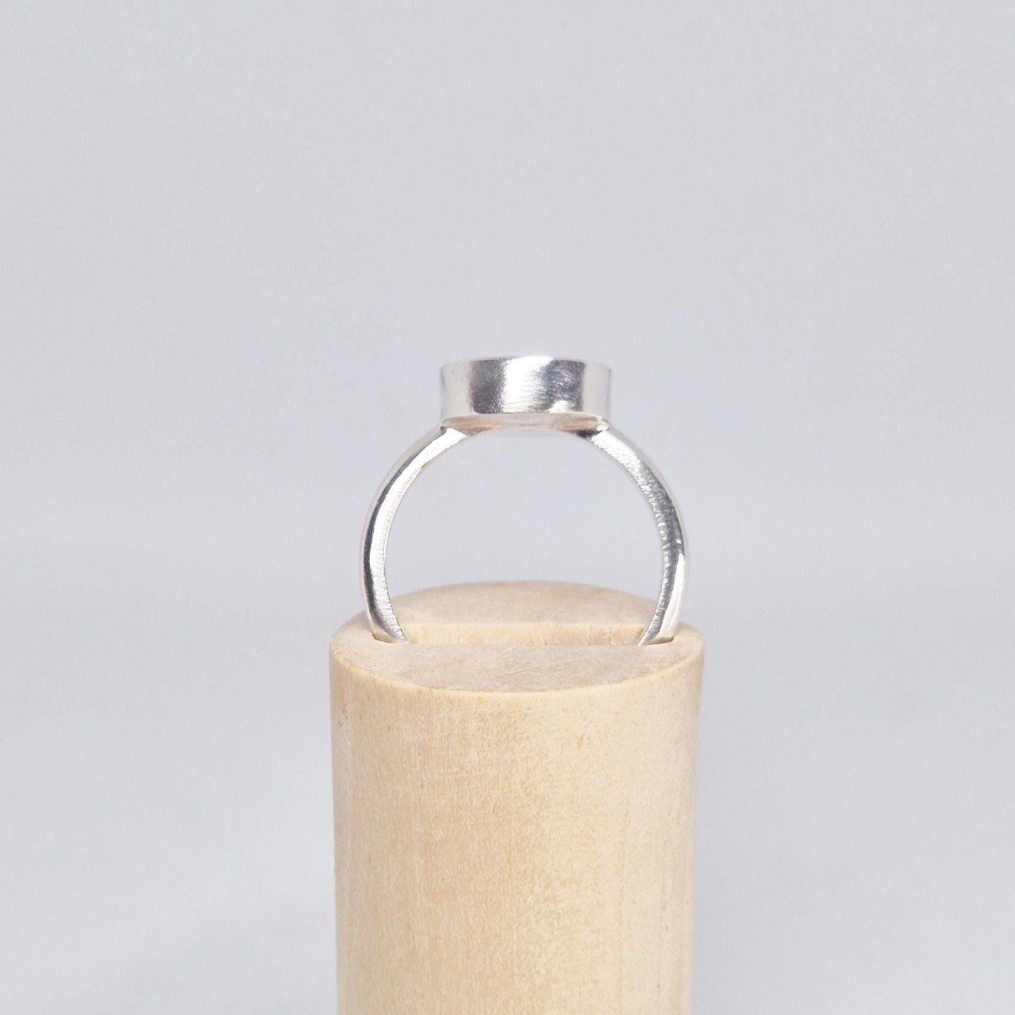 Front view of an empty sterling silver oval cabochon ring setting, standing upright on a light wood cylindrical display.