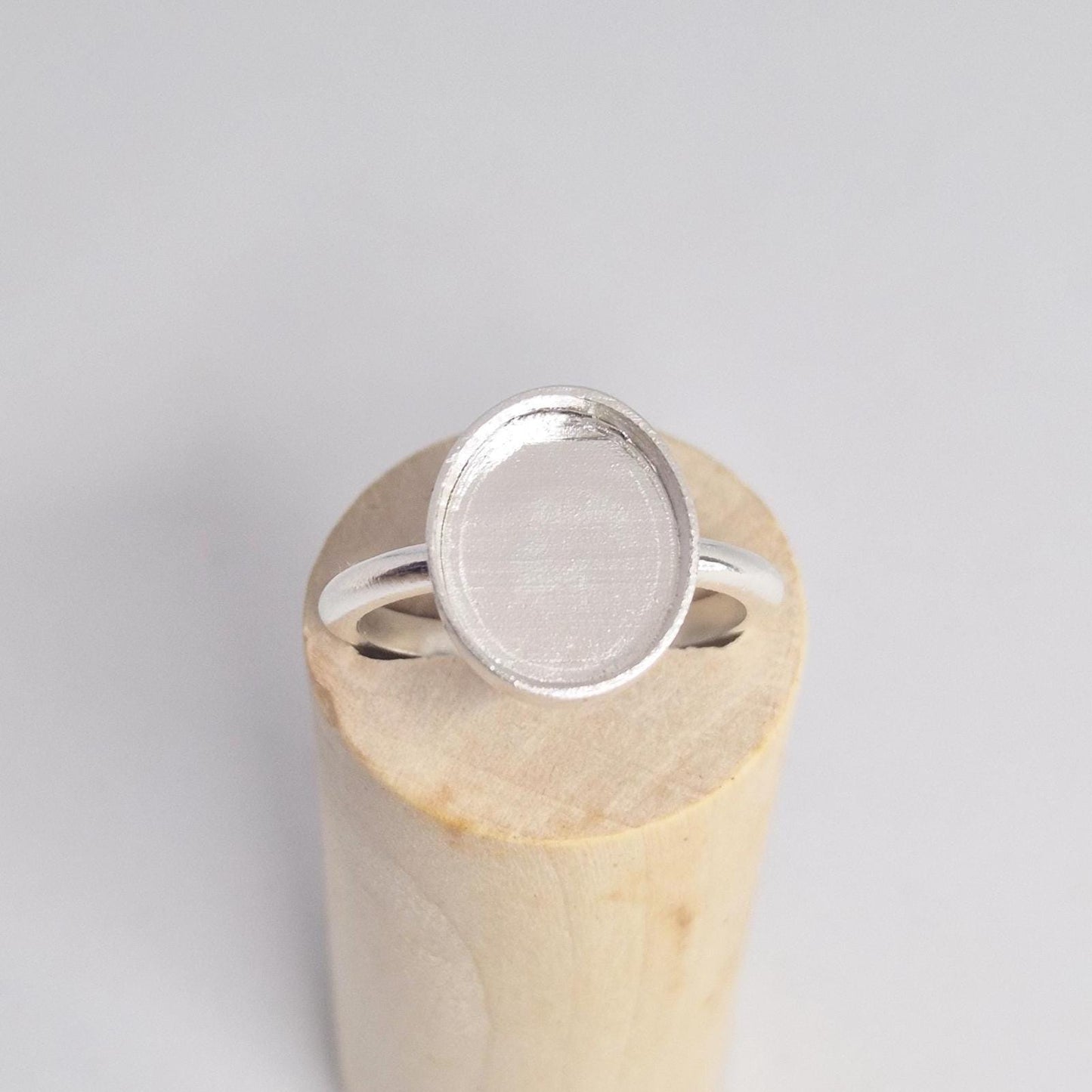 Overhead view of an empty sterling silver oval cabochon ring setting, with the band resting horizontally on a light wood cylindrical display.