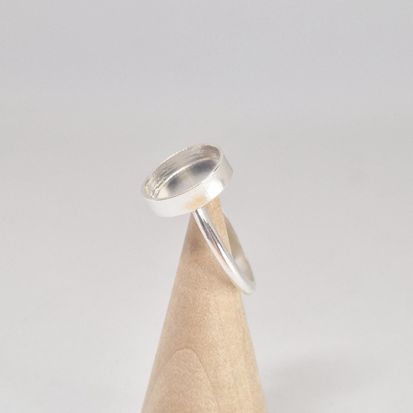 Angled side view of an empty sterling silver oval cabochon ring setting, displayed on a light wood conical stand.