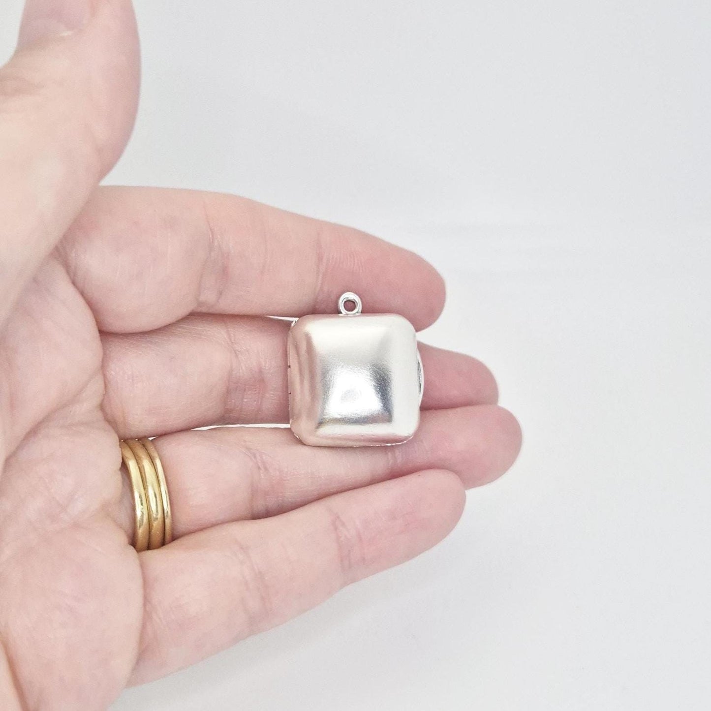 A hand holds a closed sterling silver square locket pendant, presenting its smooth, reflective surface and rounded corners from a slight angle.