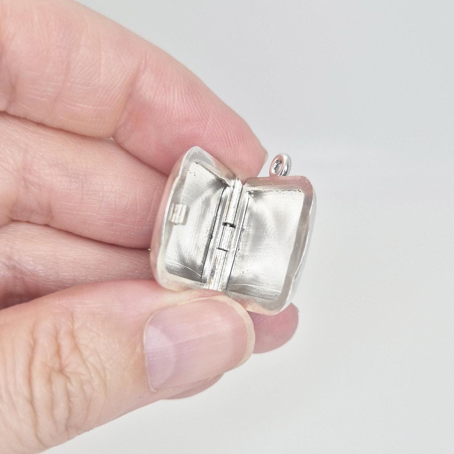 A hand holds an open sterling silver square locket pendant, allowing a clear view into its two internal compartments and the central hinge mechanism.