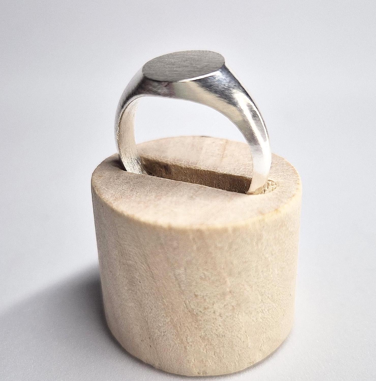 Through finger view of an oval silver signet ring in a wooden stand on a white background.