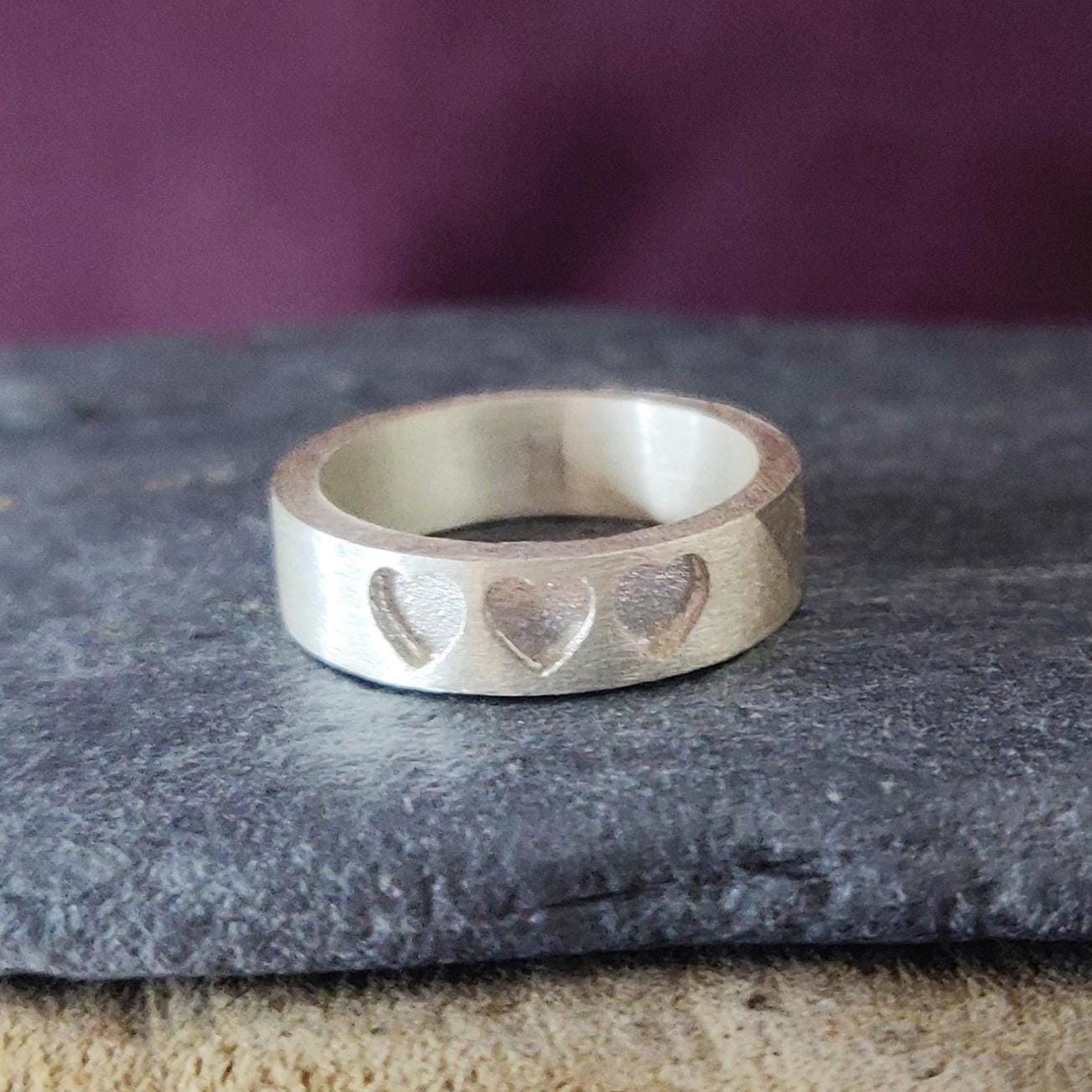 Silver memorial ring with three recessed hearts, shown on a slate stone with a purple background.