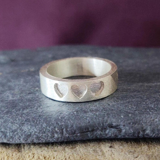 Silver memorial ring with three recessed hearts, shown on a slate stone with a purple background.