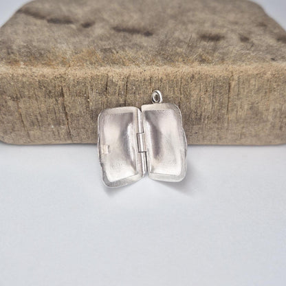 Rectangular silver locket opened on a wood block, highlighting inside surfaces and handcrafted details.