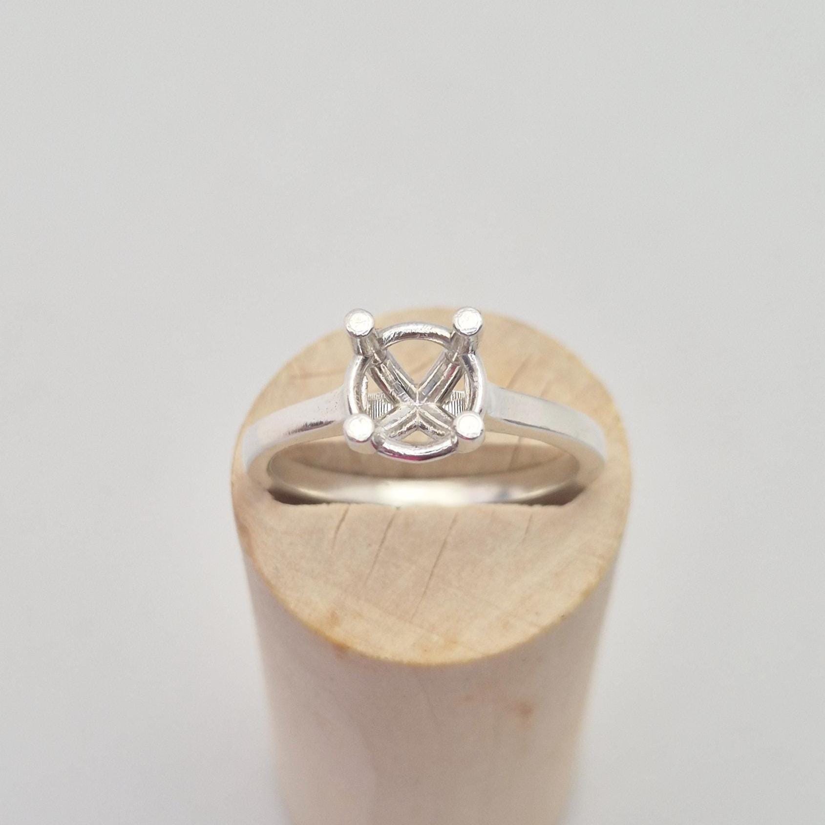 Front view of a sterling silver ring setting with a four-prong square head, centered on a cylindrical wooden stand, showcasing the clean lines of the band leading to the head.