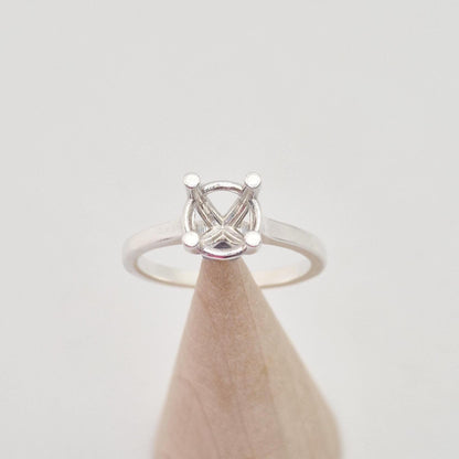 Top-down view of a sterling silver ring setting with a four-prong square head, positioned on a short, wide conical wooden stand, highlighting the symmetrical design of the prongs.
