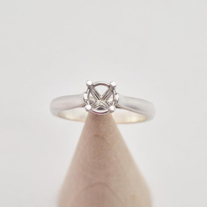Front view of a sterling silver ring setting with a four-prong round head, positioned centrally on a conical wooden stand, emphasizing the classic, simple design.