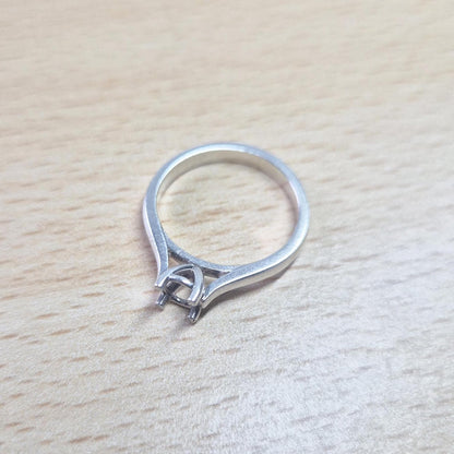 Sterling silver ring setting with a four-prong round head, resting on a light wood surface, showing the open basket-style setting and the complete ring design from above.