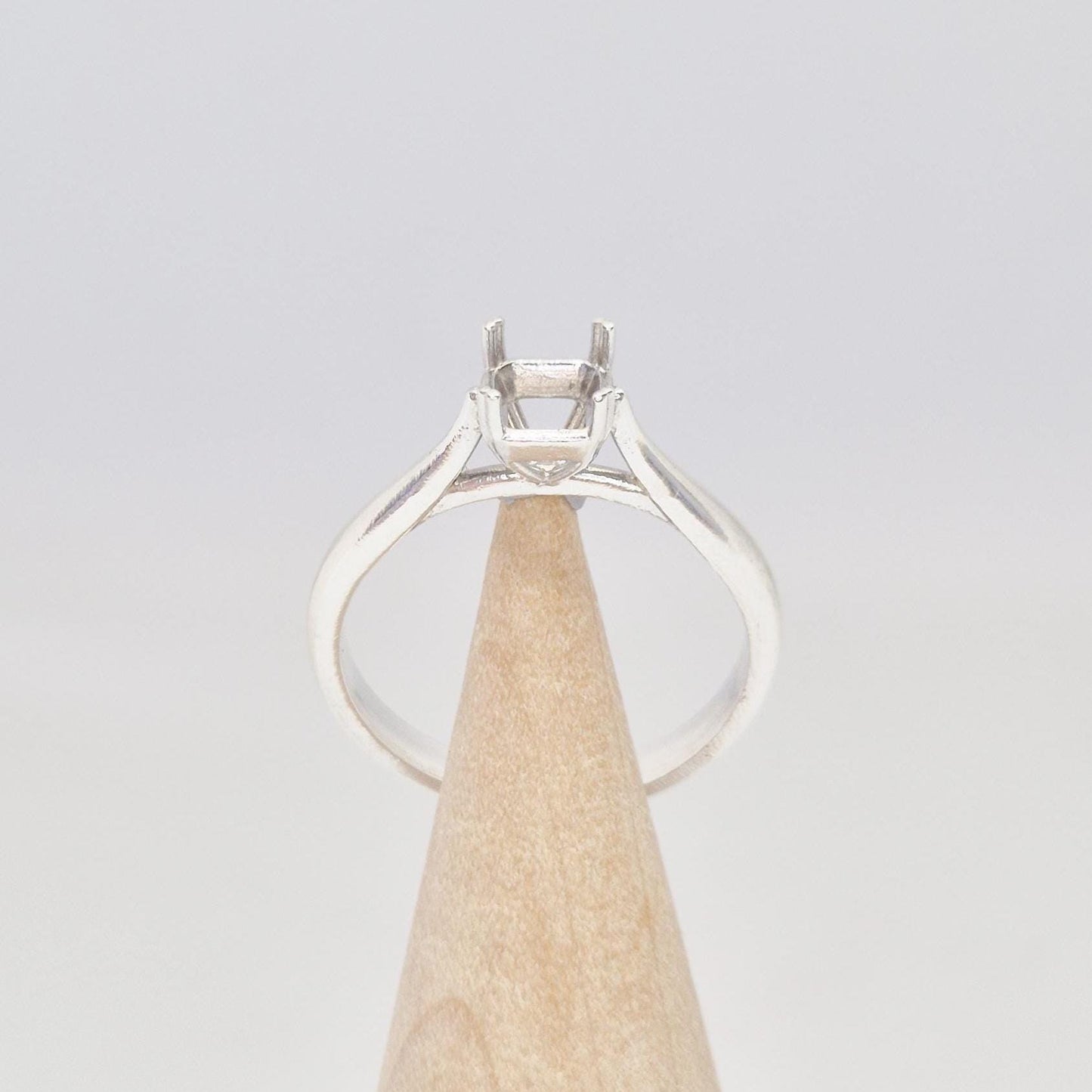 Front-facing view of a sterling silver ring setting with a four-prong princess cut head, displayed on a conical wooden stand, with the band arching elegantly towards the setting.