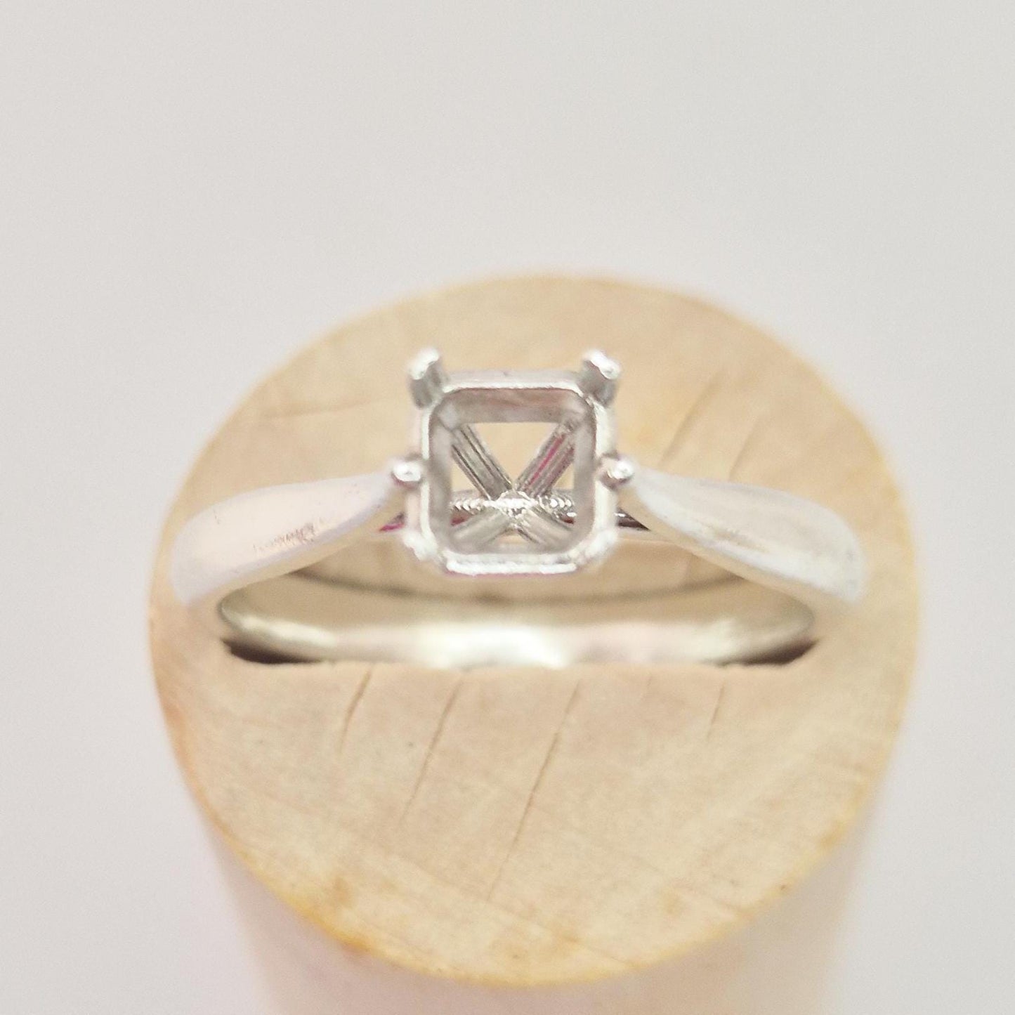 Front view of a sterling silver ring setting with a four-prong princess cut head, displayed on a cylindrical wooden stand, highlighting the symmetrical design of the setting.