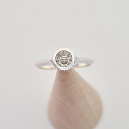 Frontal view of a silver ring setting with an empty round bezel, displayed upright on a light wooden cone. The background is bright and clear.