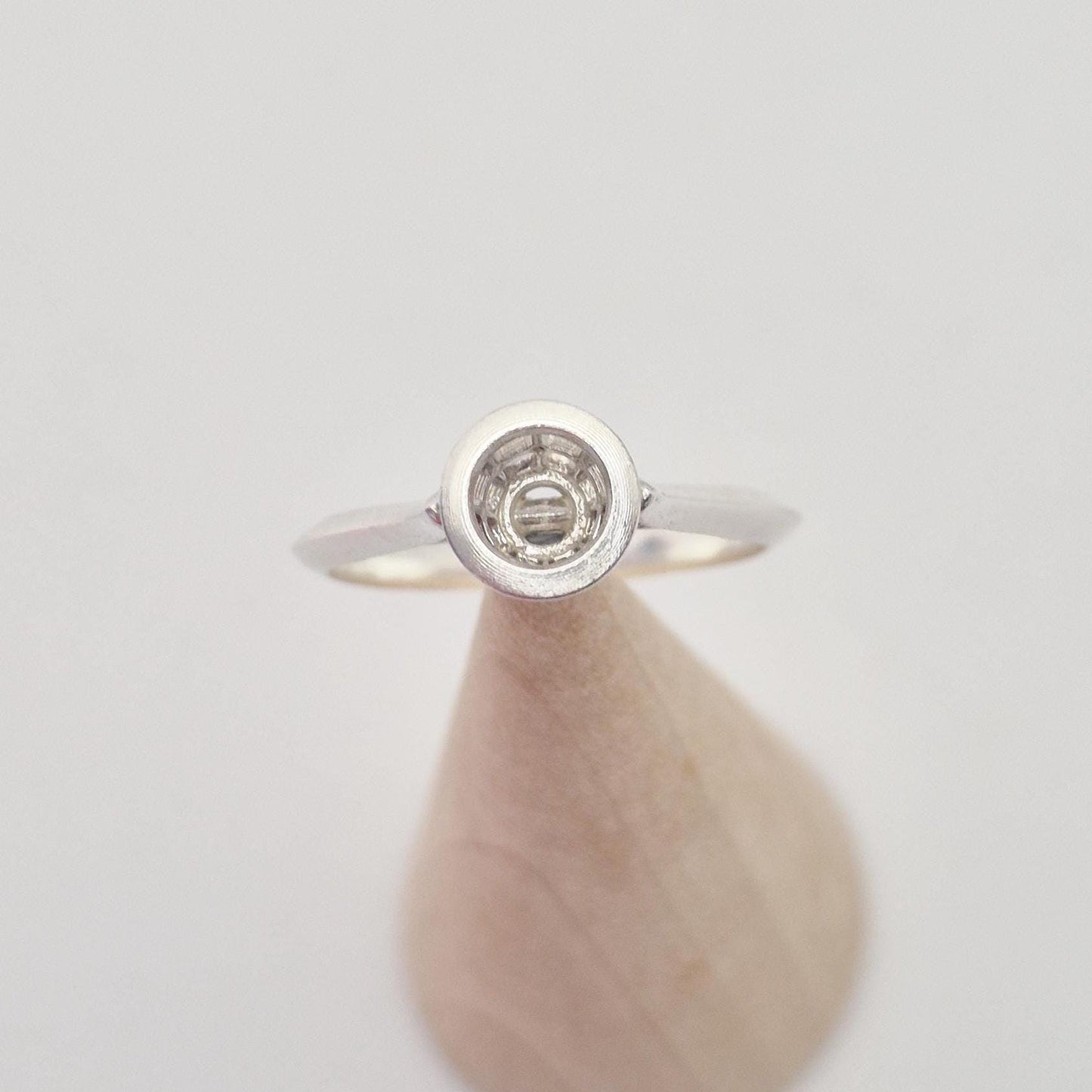 Frontal view of a silver ring setting with an empty round bezel, displayed upright on a light wooden cone. The background is bright and clear.