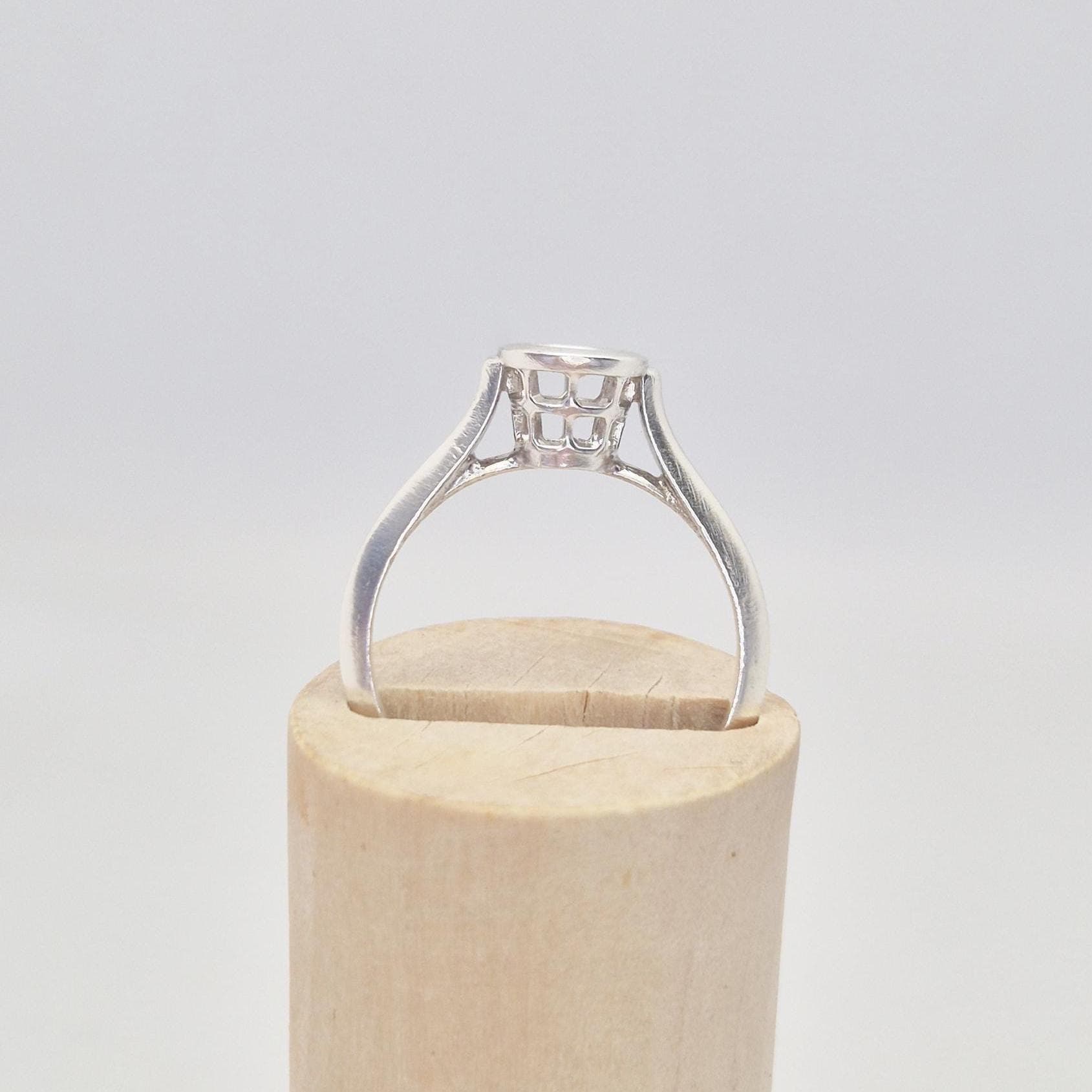 Angled side view of a silver ring setting with an empty round bezel, partially inserted into a slot on a light wooden cylinder. The details of the bezel&#39;s open basket are visible.