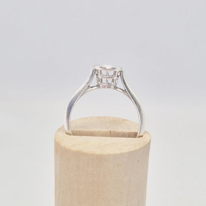 Angled side view of a silver ring setting with an empty round bezel, partially inserted into a slot on a light wooden cylinder. The details of the bezel's open basket are visible.