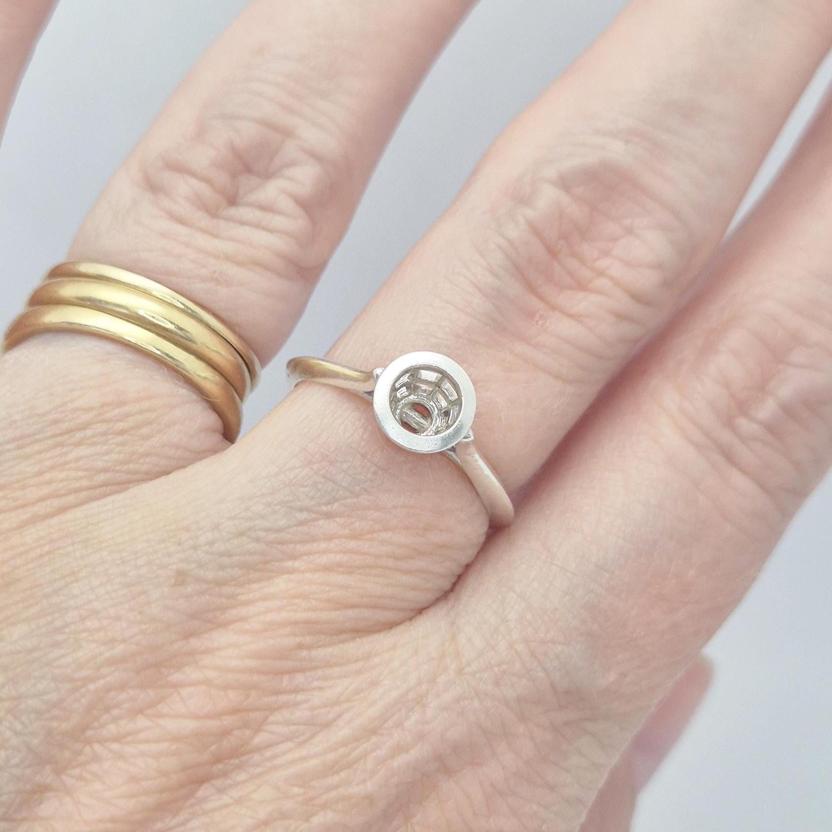 Close-up of a hand wearing a silver ring setting with a round, empty bezel, next to a stack of gold bands. The setting is designed for a round gemstone.