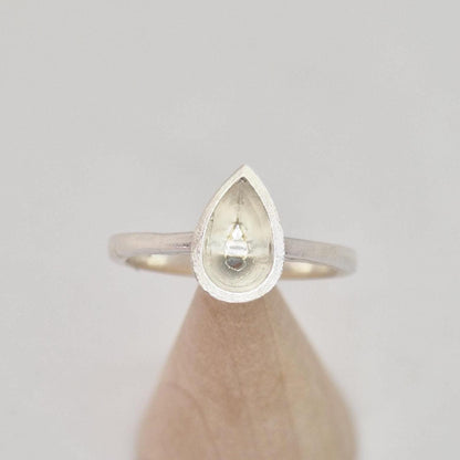 Silver pear cut bezel cathedral ring blank on a conical wooden ring holder, angled view, highlighting the empty pear-shaped bezel and textured detail on the bezel's edge.
