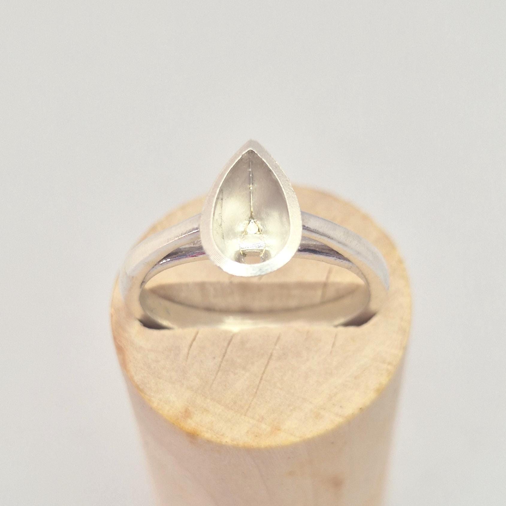 Silver pear cut bezel cathedral ring blank inserted into a cylindrical wooden ring holder, top-down view, clearly showing the empty pear-shaped bezel.