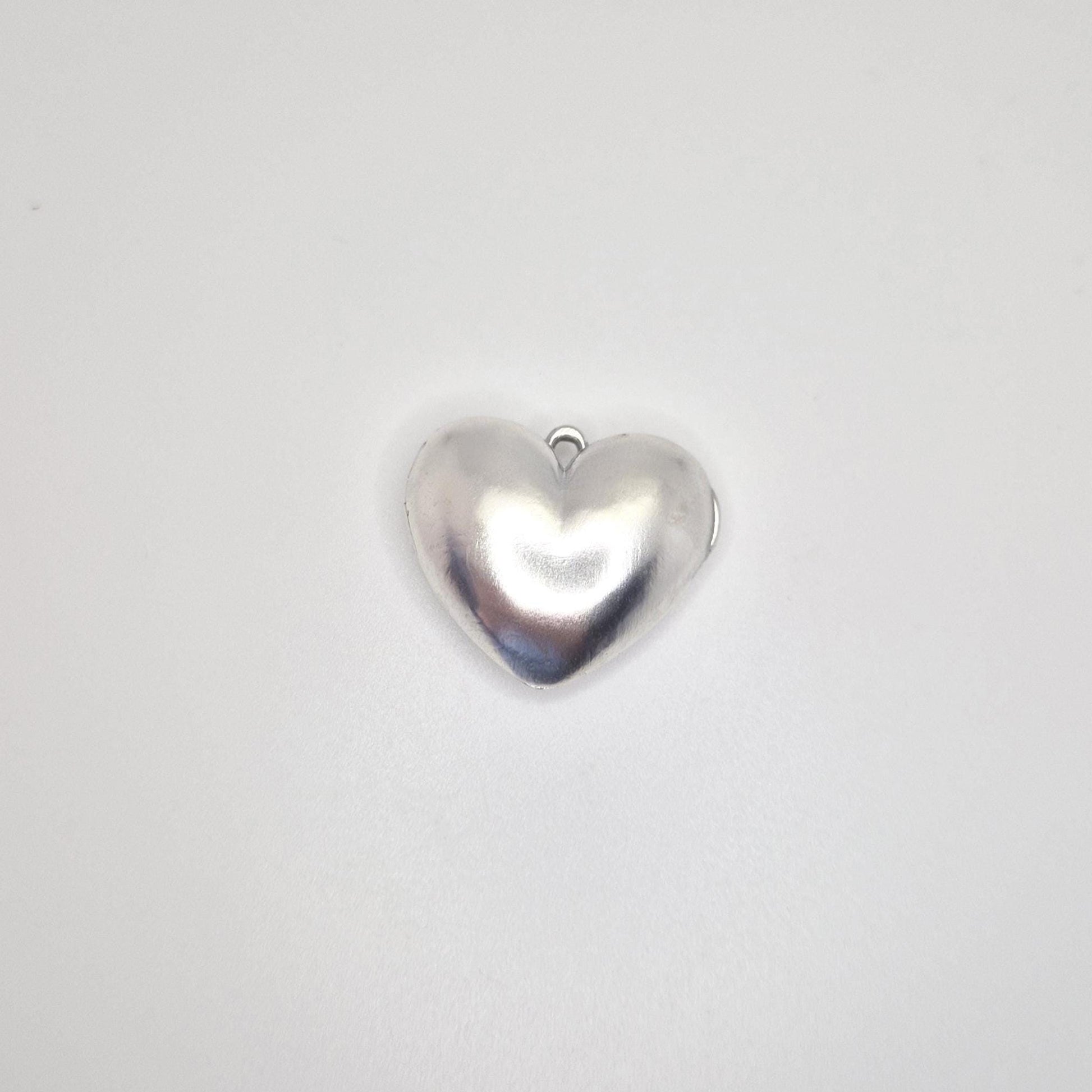 A shiny, closed, puffed silver heart locket with a small loop at the top, presented against a clean white background. The front of the locket is visible, showing its smooth, curved shape.