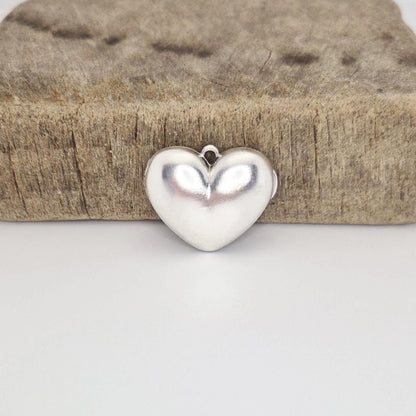 A closed, puffed silver heart locket positioned in front of a rustic wooden block on a white surface. The locket's reflective surface and the small attachment loop are clearly visible.