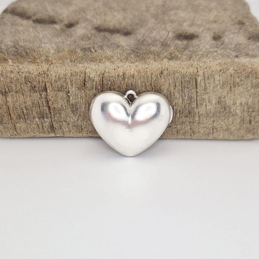 A closed, puffed silver heart locket positioned in front of a rustic wooden block on a white surface. The locket&#39;s reflective surface and the small attachment loop are clearly visible.