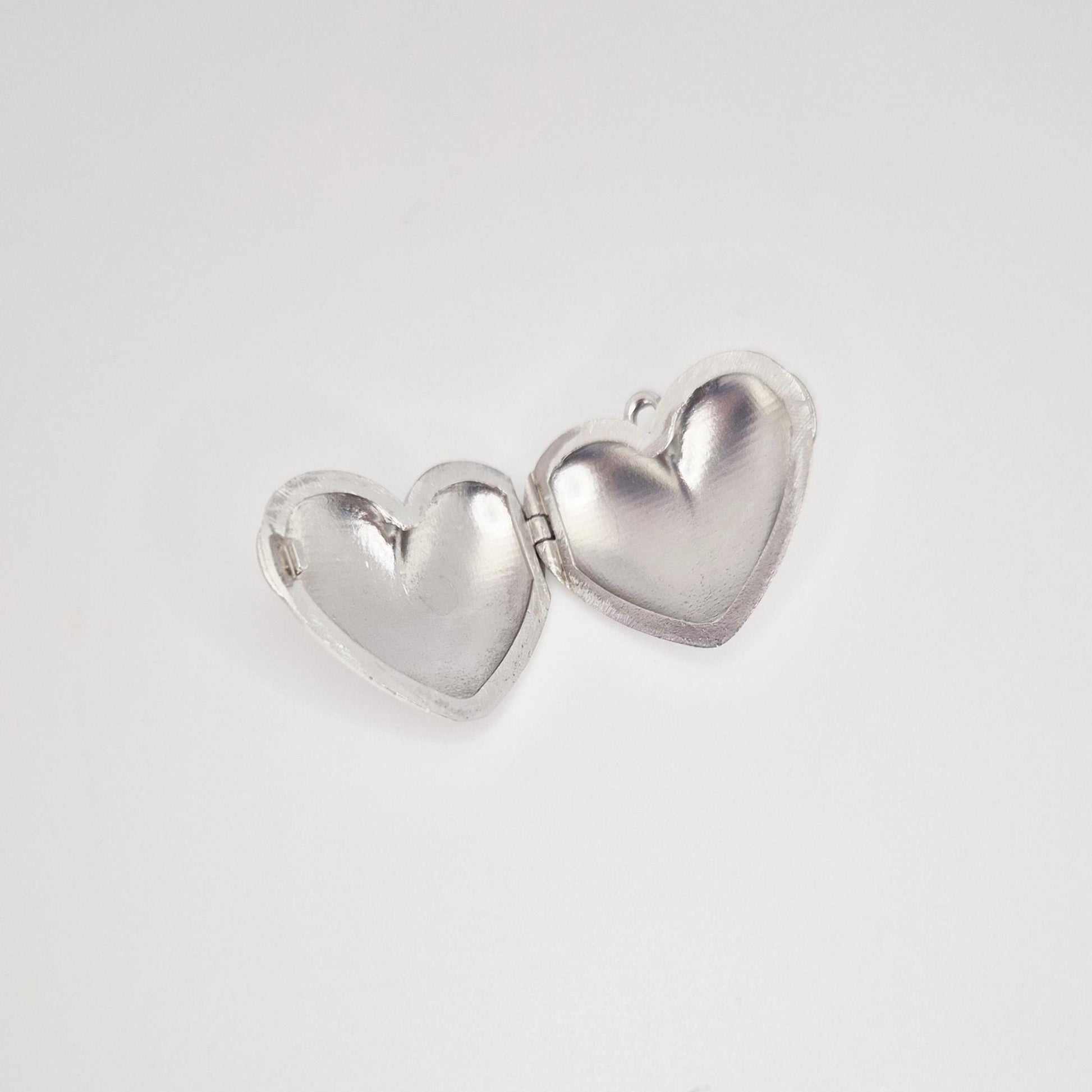 An open, puffed silver heart locket against a plain white background. The two halves of the heart are spread open, showing the smooth, concave interior of each side, with a small jump ring visible on the right half.