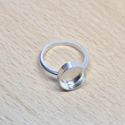 Top-down view of a sterling silver oval cabochon ring setting resting on a light wood surface, showcasing the empty bezel.