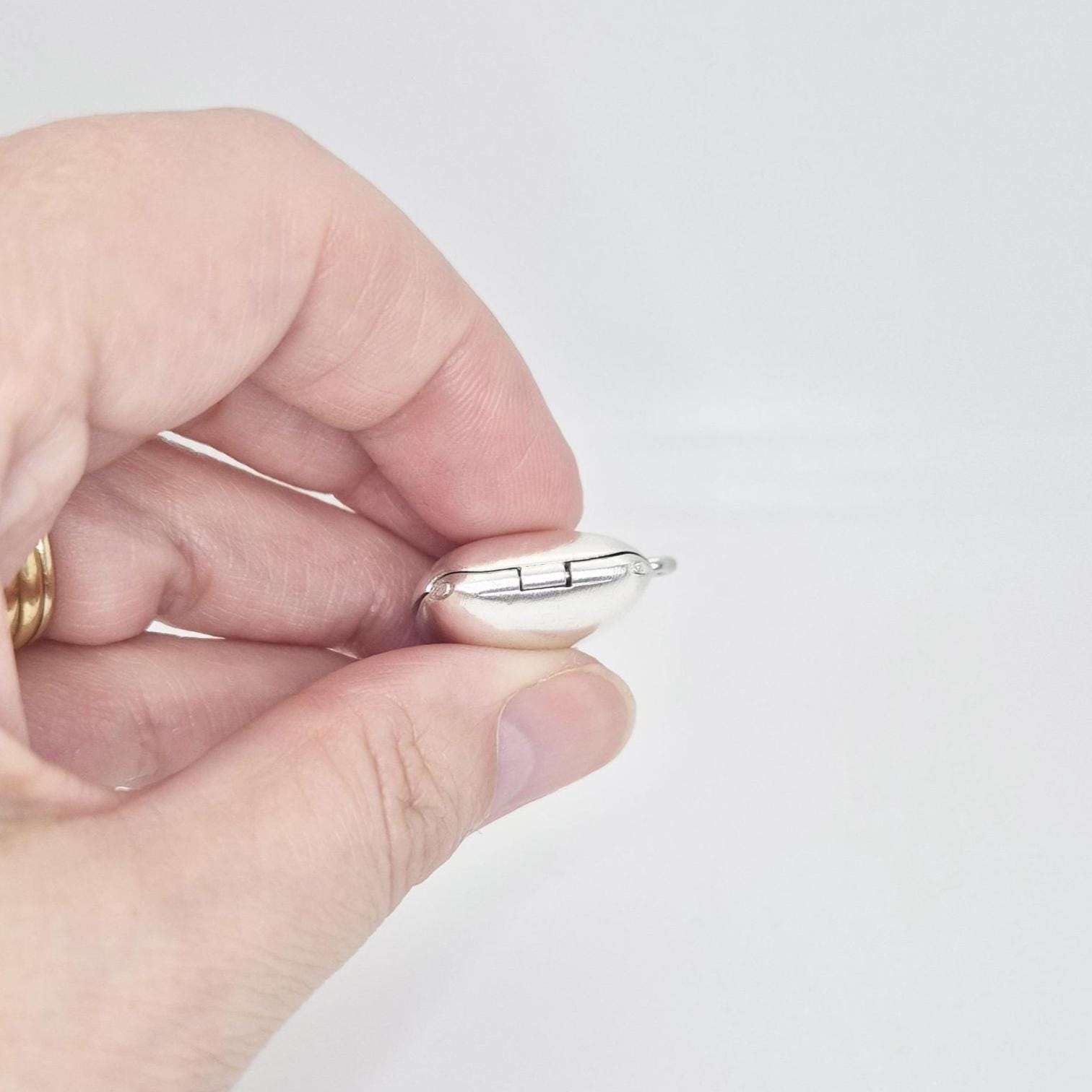 A hand holds a polished sterling silver square locket pendant, showing its hinged closure and the rounded edges from a side view.